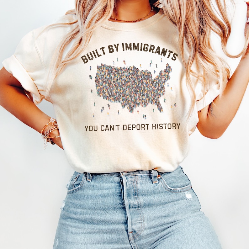 May include: Cream-colored t-shirt with the text "BUILT BY IMMIGRANTS" above a graphic of the United States filled with tiny figures. Below the graphic, the text reads "YOU CAN'T DEPORT HISTORY". The shirt is paired with blue jeans.