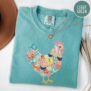 Op de afbeelding: Een lichtblauwgroen Comfort Colors T-shirt met een geborduurd kippenontwerp. De kip is gemaakt van kleurrijke stoffen patches met patronen zoals ruitjes, aardbeien en ananassen. Een gouden ketting met een hanger ligt op het shirt.