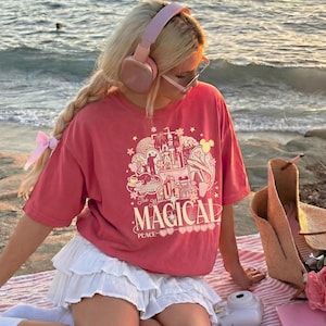 May include: A coral-colored short-sleeve t-shirt featuring a graphic print of a castle, stars, and the text The Most MAGICAL PLACE. The shirt is paired with a white tiered mini skirt, light pink over-ear headphones, and clear-framed glasses.