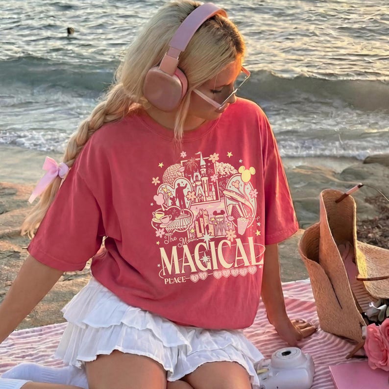 May include: A coral-colored short-sleeve t-shirt featuring a graphic print of a castle, stars, and the text The Most MAGICAL PLACE. The shirt is paired with a white tiered mini skirt, light pink over-ear headphones, and clear-framed glasses.