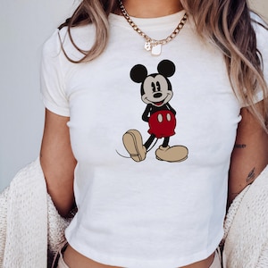May include: White crop top featuring a full-body illustration of Mickey Mouse. The cartoon character is depicted in his classic outfit: black shorts, red pants, and large yellow shoes. The top has short sleeves and a round neckline.