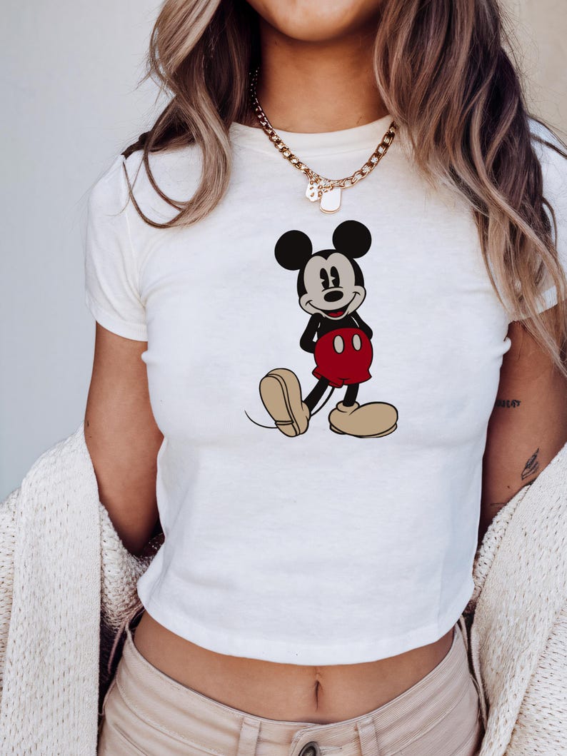 May include: White crop top featuring a full-body illustration of Mickey Mouse. The cartoon character is depicted in his classic outfit: black shorts, red pants, and large yellow shoes. The top has short sleeves and a round neckline.