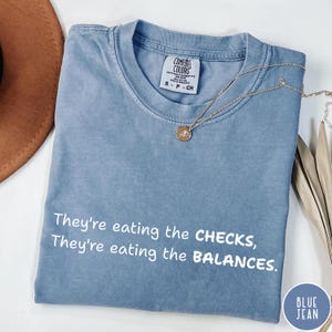 Puede incluir: Una camiseta azul claro con el texto "They're eating the CHECKS, They're eating the BALANCES." impreso en blanco. La camiseta está hecha de un material suave y cómodo y tiene un aspecto vintage.