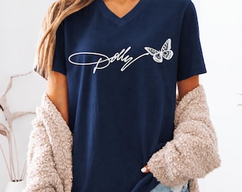 May include: A navy blue V-neck t-shirt featuring the white cursive text Dolly followed by a white butterfly graphic. The shirt has short sleeves and a standard fit, providing a clean look with a prominent white design centered on the chest area.