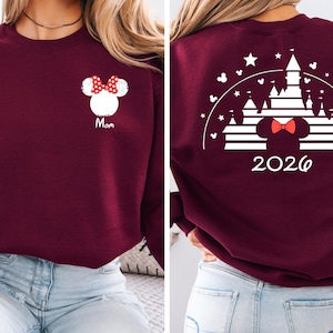Puede incluir: Sudadera burdeos con un dise&ntilde;o de Minnie Mouse blanco y un lazo rojo, con la palabra "Mom". La parte trasera presenta un dise&ntilde;o de castillo blanco, un lazo de Minnie Mouse y el a&ntilde;o "2026".