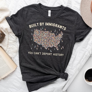 May include: Dark gray t-shirt with the text "BUILT BY IMMIGRANTS YOU CAN'T DEPORT HISTORY" and a graphic of the United States filled with diverse figures. The shirt is styled with a hat, a book, a clock, and a cup of coffee.