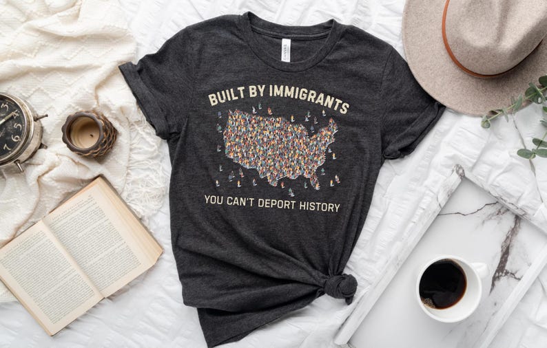 May include: Dark gray t-shirt with the text "BUILT BY IMMIGRANTS YOU CAN'T DEPORT HISTORY" and a graphic of the United States filled with diverse figures. The shirt is styled with a hat, a book, a clock, and a cup of coffee.