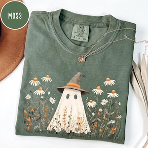 Puede incluir: Camiseta verde musgo con un fantasma con sombrero de bruja, rodeado de margaritas y flores silvestres. La camiseta tiene cuello redondo y mangas cortas. Un collar plateado con un colgante está sobre la camiseta.