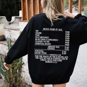 May include: Black sweatshirt with white text that reads "JESUS PAID IT ALL" and a receipt-style list of sins and the price paid for them. The text at the bottom reads "FOR THE WAGES OF SIN IS DEATH, BUT THE GIFT OF GOD IS ETERNAL LIFE IN CHRIST JESUS OUR LORD. ROMANS 6.23"