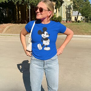 May include: A royal blue t-shirt featuring a classic Mickey Mouse graphic. The shirt is paired with light blue jeans and a white purse. The person is wearing sunglasses and gold hoop earrings.
