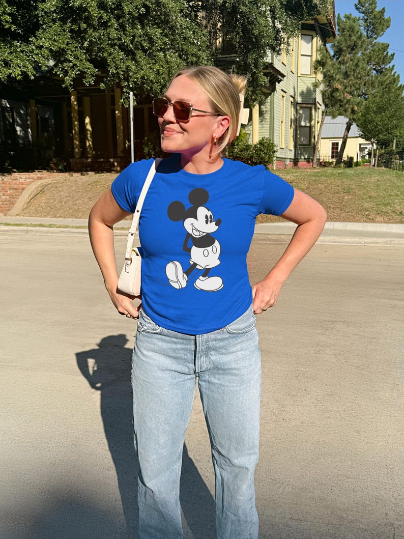 May include: A royal blue t-shirt featuring a classic Mickey Mouse graphic. The shirt is paired with light blue jeans and a white purse. The person is wearing sunglasses and gold hoop earrings.
