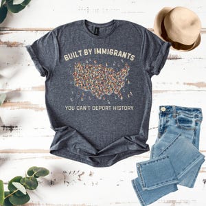 May include: Dark gray t-shirt with the text "BUILT BY IMMIGRANTS" above a graphic of the United States filled with diverse figures. Below, the text reads "YOU CAN'T DEPORT HISTORY." A straw hat and blue jeans are also in the image.
