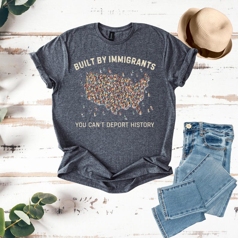 May include: Dark gray t-shirt with the text "BUILT BY IMMIGRANTS" above a graphic of the United States filled with diverse figures. Below, the text reads "YOU CAN'T DEPORT HISTORY." A straw hat and blue jeans are also in the image.