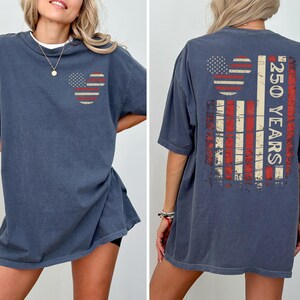 May include: A navy blue t-shirt with a distressed American flag design. The front features a Mickey Mouse head shape with the flag's stars and stripes. The back displays a larger flag design with the text "250 YEARS".