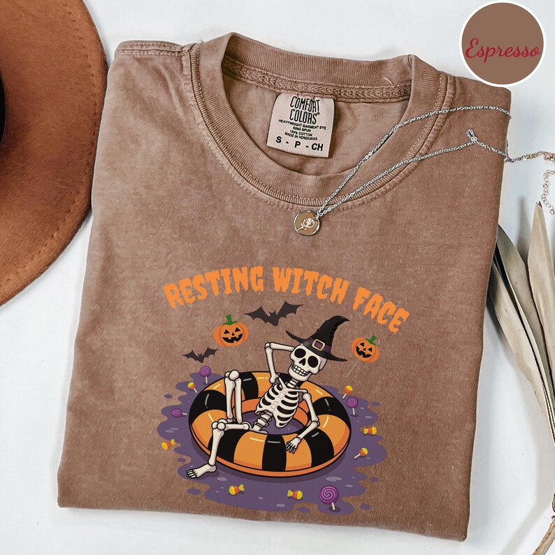 May include: Espresso-colored t-shirt with the phrase "RESTING WITCH FACE" in orange. The graphic features a skeleton in a witch hat relaxing in a black and orange inner tube, with pumpkins, bats, and candy. A silver necklace is draped across the shirt.