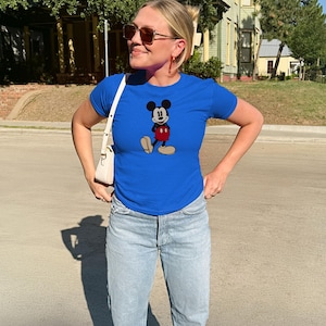 May include: A royal blue t-shirt featuring a classic Mickey Mouse graphic. The shirt is paired with light wash blue jeans. The person is wearing sunglasses and a white purse.