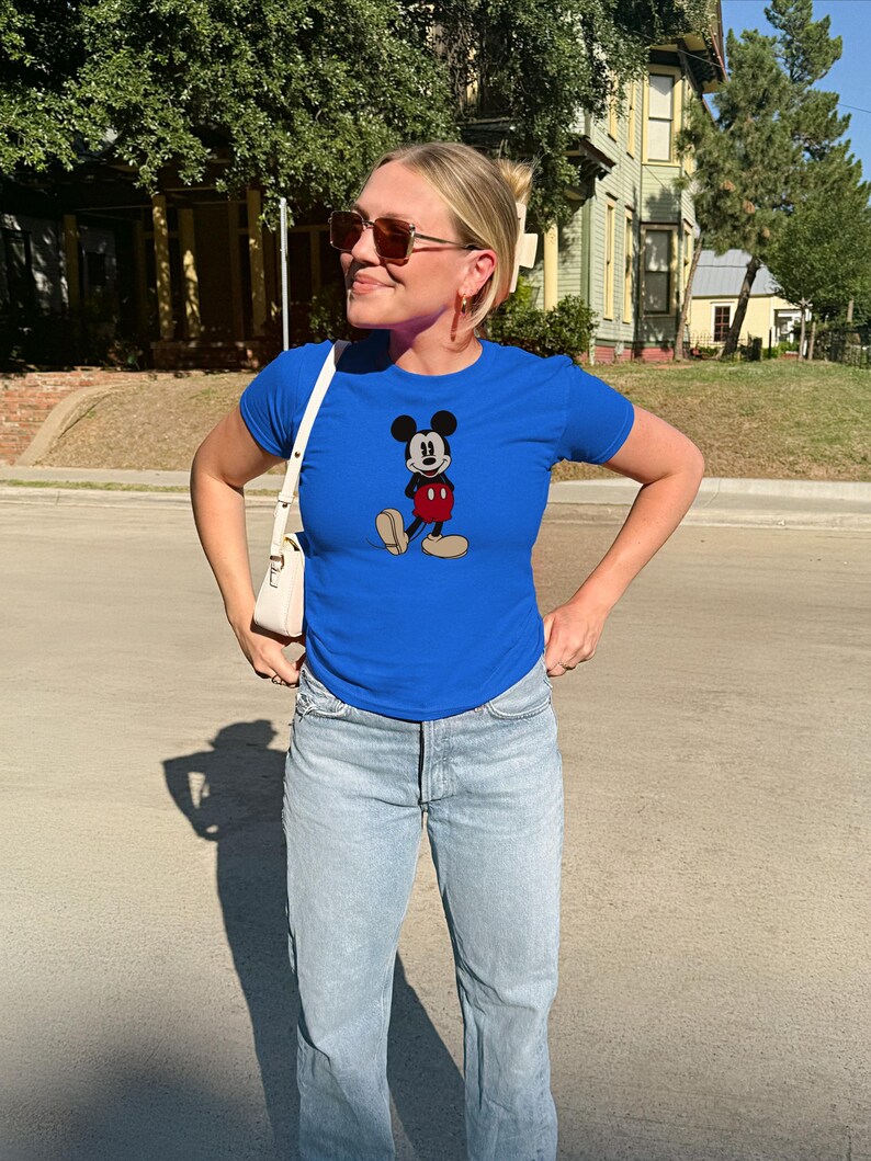May include: A royal blue t-shirt featuring a classic Mickey Mouse graphic. The shirt is paired with light wash blue jeans. The person is wearing sunglasses and a white purse.