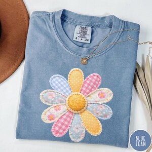 May include: A blue Comfort Colors t-shirt with a large, colorful daisy applique. The daisy features petals in gingham, floral, and polka dot patterns. A gold necklace with a pendant rests on the shirt. The shirt is labeled "Blue Jean".