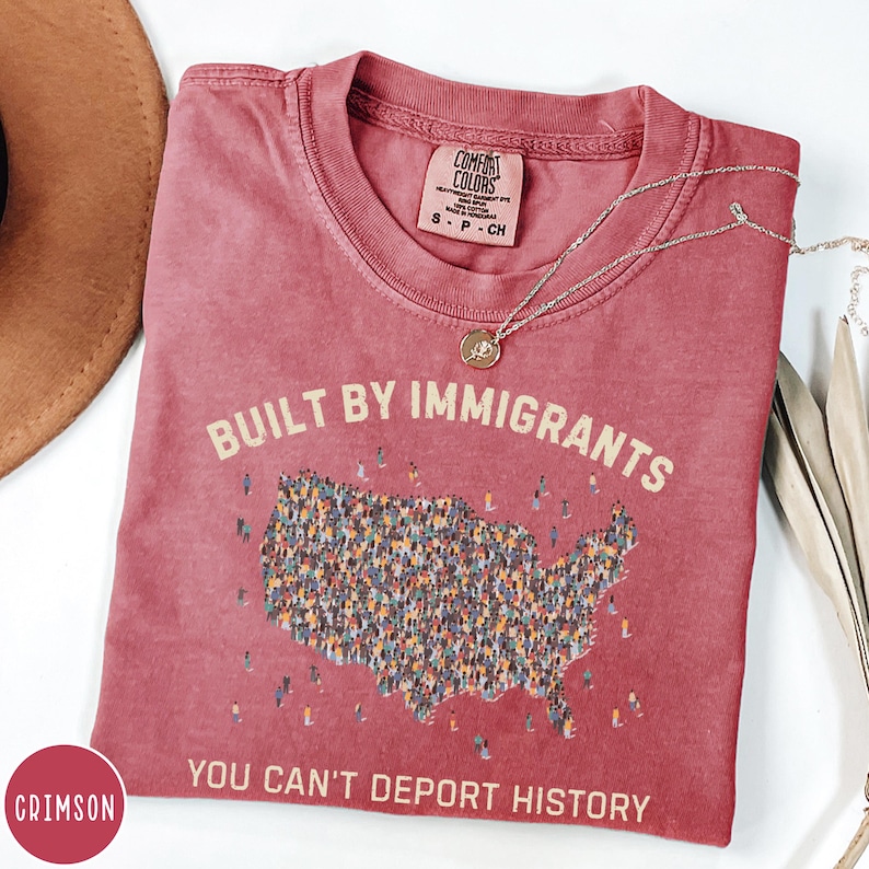 May include: Crimson-colored t-shirt with the text "BUILT BY IMMIGRANTS" above a map of the United States filled with tiny figures. The phrase "YOU CAN'T DEPORT HISTORY" is printed below the map. A silver necklace with a pendant rests on the shirt.
