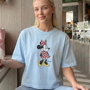 May include: A light blue t-shirt featuring a Minnie Mouse graphic. The cartoon character is wearing a red polka dot dress, bow, and yellow shoes. The shirt is part of a casual wear collection.