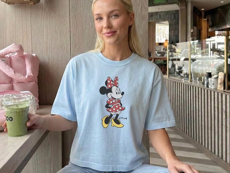 May include: A light blue t-shirt featuring a Minnie Mouse graphic. The cartoon character is wearing a red polka dot dress, bow, and yellow shoes. The shirt is part of a casual wear collection.