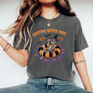 May include: Dark gray t-shirt with the text "RESTING WITCH FACE" above a skeleton wearing a witch hat, relaxing on a black and orange inflatable ring. The design includes pumpkins, bats, and candy.