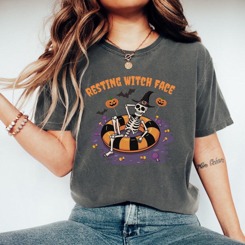 May include: Dark gray t-shirt with the text "RESTING WITCH FACE" above a skeleton wearing a witch hat, relaxing on a black and orange inflatable ring. The design includes pumpkins, bats, and candy.