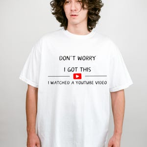 May include: White t-shirt with the text "DON'T WORRY I GOT THIS" above a YouTube play button and the phrase "I WATCHED A YOUTUBE VIDEO". The shirt is a casual, short-sleeved style.