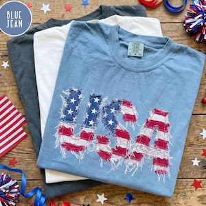 May include: Three t-shirts in blue, white, and gray are stacked on a wooden surface. The top shirt is light blue and features the word "USA" in a distressed American flag design. Patriotic stars and decorations surround the shirts.