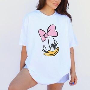 May include: White t-shirt featuring a cartoon graphic of Daisy Duck's face. The design includes a pink bow, black eyes and eyelashes, and a yellow beak. The shirt is a casual, oversized fit.