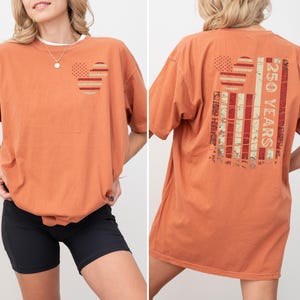 May include: Burnt orange t-shirt with a graphic design on the front and back. The front features two circles with a dotted and striped pattern. The back displays a distressed American flag design with the text "250 YEARS".