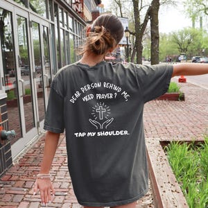 May include: A person wearing a dark gray t-shirt with the text "Dear person behind me, need prayer? Tap my shoulder." and a white cross graphic.