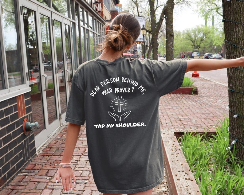 May include: A person wearing a dark gray t-shirt with the text "Dear person behind me, need prayer? Tap my shoulder." and a white cross graphic.