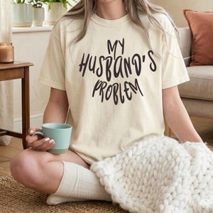 May include: Cream-colored t-shirt with the text "MY HUSBAND'S PROBLEM" in black script. The shirt is worn by a person sitting cross-legged, holding a blue mug. A white knitted blanket is in the foreground.