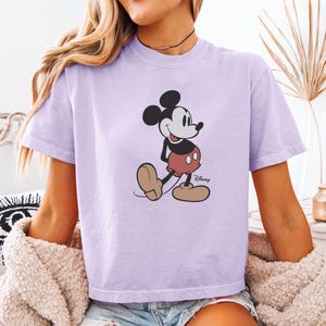 以下が含まれることがあります： ミッキーマウスのグラフィックが描かれた薄紫色のTシャツ。漫画のキャラクターは、黒い耳、白い顔、赤いショートパンツ、大きな茶色の靴を履いた、彼の象徴的なポーズで描かれています。ディズニーのロゴが見えます。