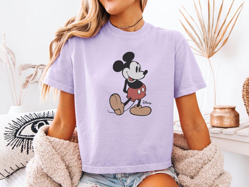 以下が含まれることがあります： ミッキーマウスのグラフィックが描かれた薄紫色のTシャツ。漫画のキャラクターは、黒い耳、白い顔、赤いショートパンツ、大きな茶色の靴を履いた、彼の象徴的なポーズで描かれています。ディズニーのロゴが見えます。
