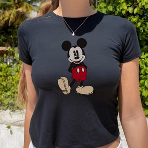 May include: Black crop top featuring a classic Mickey Mouse graphic. The cartoon character is depicted in his iconic pose, wearing red shorts and beige shoes. The top is a fitted style, perfect for casual wear.