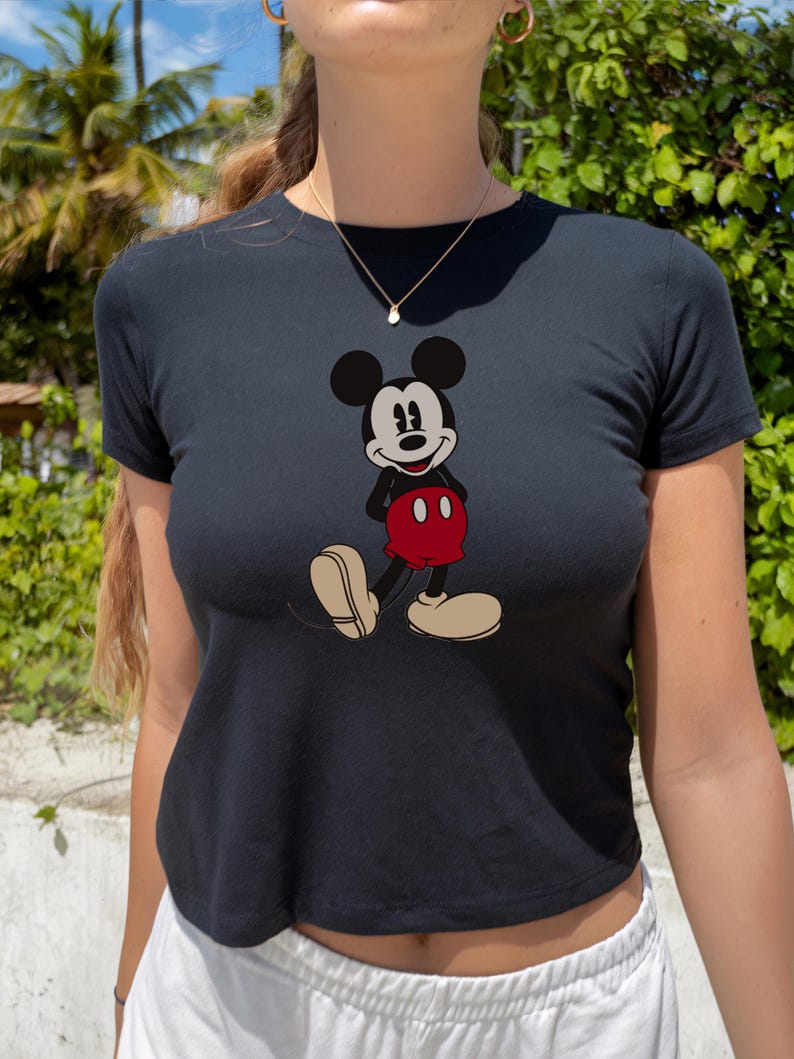 May include: Black crop top featuring a classic Mickey Mouse graphic. The cartoon character is depicted in his iconic pose, wearing red shorts and beige shoes. The top is a fitted style, perfect for casual wear.