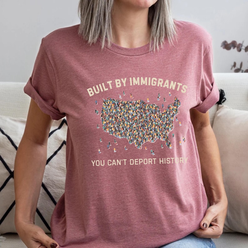 May include: A dusty rose t-shirt with the text "BUILT BY IMMIGRANTS" above a map of the United States filled with diverse figures. Below the map, the text reads "YOU CAN'T DEPORT HISTORY".