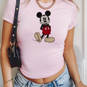 May include: A light pink t-shirt featuring a graphic of Mickey Mouse in his classic pose. The cartoon character is depicted in black, red, and beige. The shirt has short sleeves.