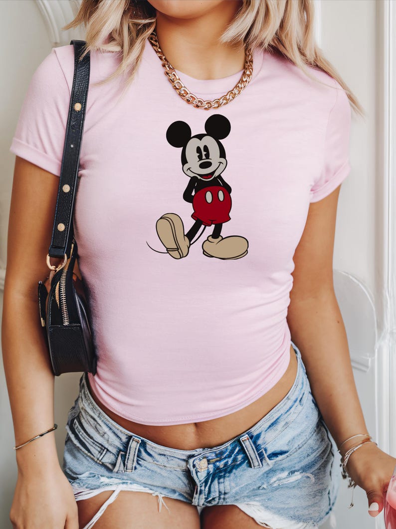 May include: A light pink t-shirt featuring a graphic of Mickey Mouse in his classic pose. The cartoon character is depicted in black, red, and beige. The shirt has short sleeves.