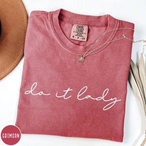 Puede incluir: Camiseta de color carmes&iacute; con la frase "do it lady" en escritura blanca. La camiseta tiene cuello redondo y una etiqueta que dice "Comfort Colors". Un collar plateado con un colgante est&aacute; drapeado sobre la camiseta. Un sombrero marr&oacute;n es parcialmente visible.