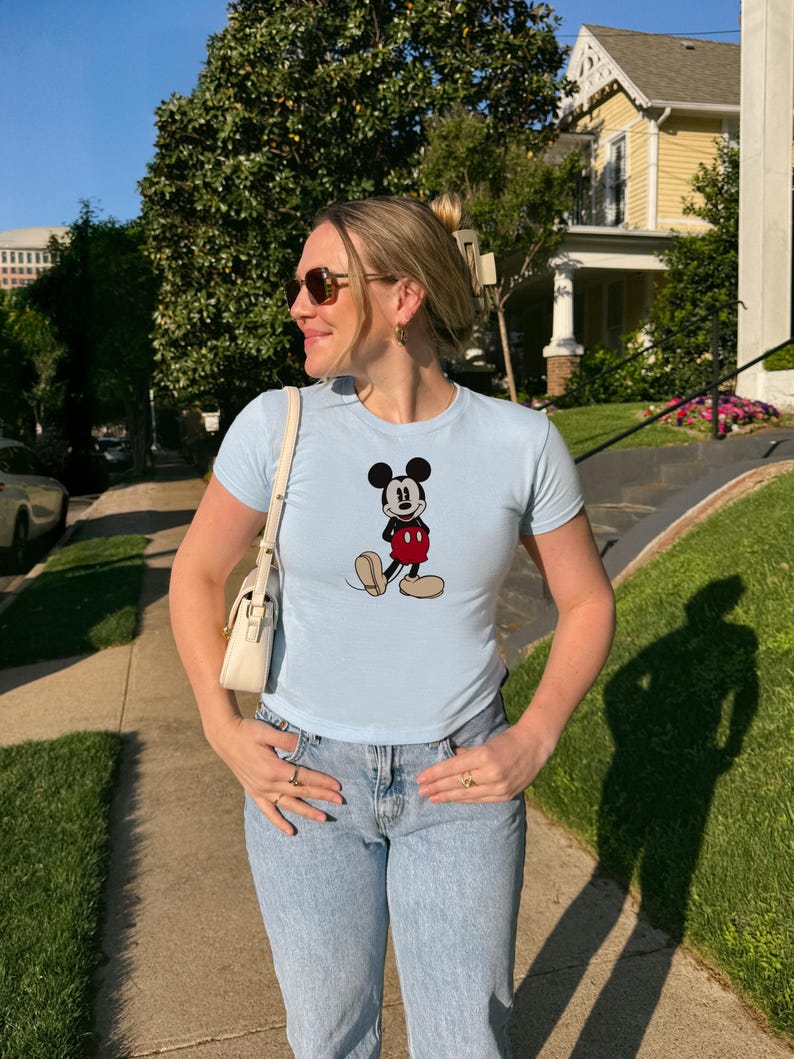May include: A light blue t-shirt featuring a classic Mickey Mouse graphic. The shirt has short sleeves and is paired with light wash blue jeans. The person is wearing sunglasses and carrying a white purse.
