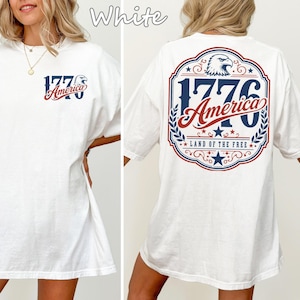 May include: White t-shirt with a patriotic design. The front features "1776 America" in blue and red. The back displays a larger design with an eagle, the number "1776", and the words "America" and "Land of the Free" in a similar color scheme.