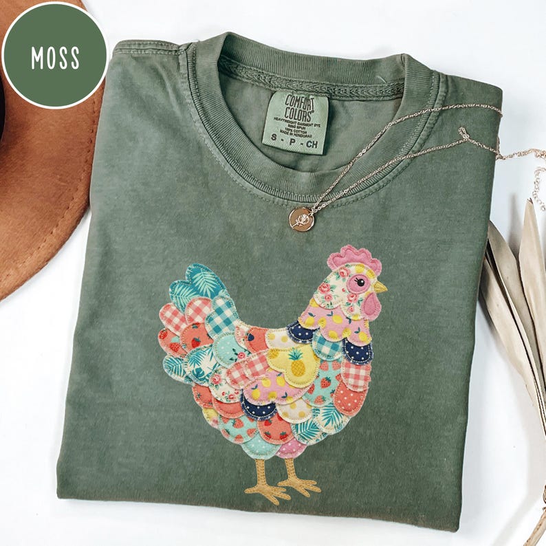 Op de afbeelding: Een mosgroen Comfort Colors T-shirt met een kleurrijk patchwork kippenontwerp. De kip heeft een roze kam, een gele snavel en is versierd met verschillende stoffen met patronen. Een zilveren ketting met een hanger is over het shirt gedrapeerd.