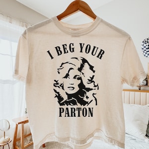 Könnte beinhalten: Ein cremefarbenes Kurzarm-T-Shirt mit einem schwarzen Grafikdruck einer Frau mit voluminösem Haar. Der Text "I BEG YOUR" ist in Schwarz über der Grafik gedruckt, und das Wort "PARTON" ist in Schwarz unter der Grafik gedruckt.