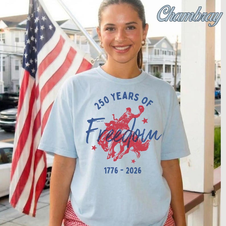 May include: Light blue t-shirt with the text "250 YEARS OF Freedom 1776-2026" and a red graphic of a person riding a horse. An American flag is in the background.