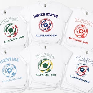 May include: White t-shirts featuring soccer ball designs with country names and "ALL FOR ONE - 2026". Countries include Mexico, United States, Canada, Argentina, Brazil, and France. Each ball has a unique color scheme.