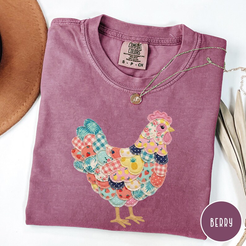 Op de afbeelding: Berry-kleurig Comfort Colors T-shirt met een patchwork kippenontwerp. De kip heeft verschillende stoffen met patronen in blauw, roze, geel en rood. Een gouden ketting met een hanger ligt op het shirt. Het shirt is gelabeld S-P-CH.