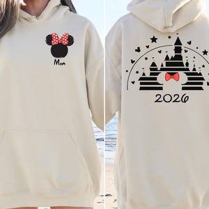 Puede incluir: Sudadera con capucha color crema con bolsillo delantero y capucha. La parte delantera presenta una cabeza de Minnie Mouse negra con un lazo de lunares rojos y blancos y la palabra "Mom" en escritura negra. La parte trasera muestra un dise&ntilde;o de castillo con un lazo rojo y el a&ntilde;o "2026".